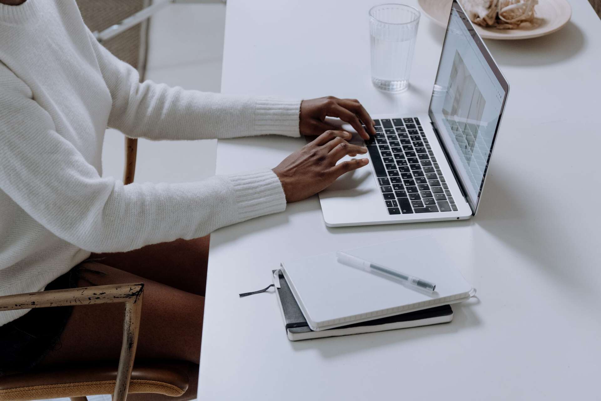 person writing on a laptop, pen and paper at her side