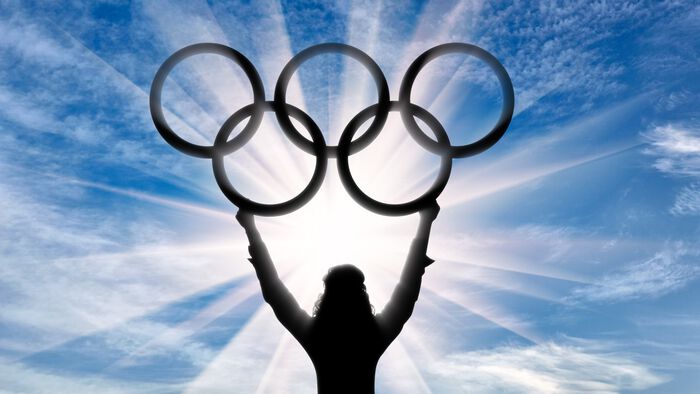 A woman in silhouette holds the 5 Olympic rings up to the sky.