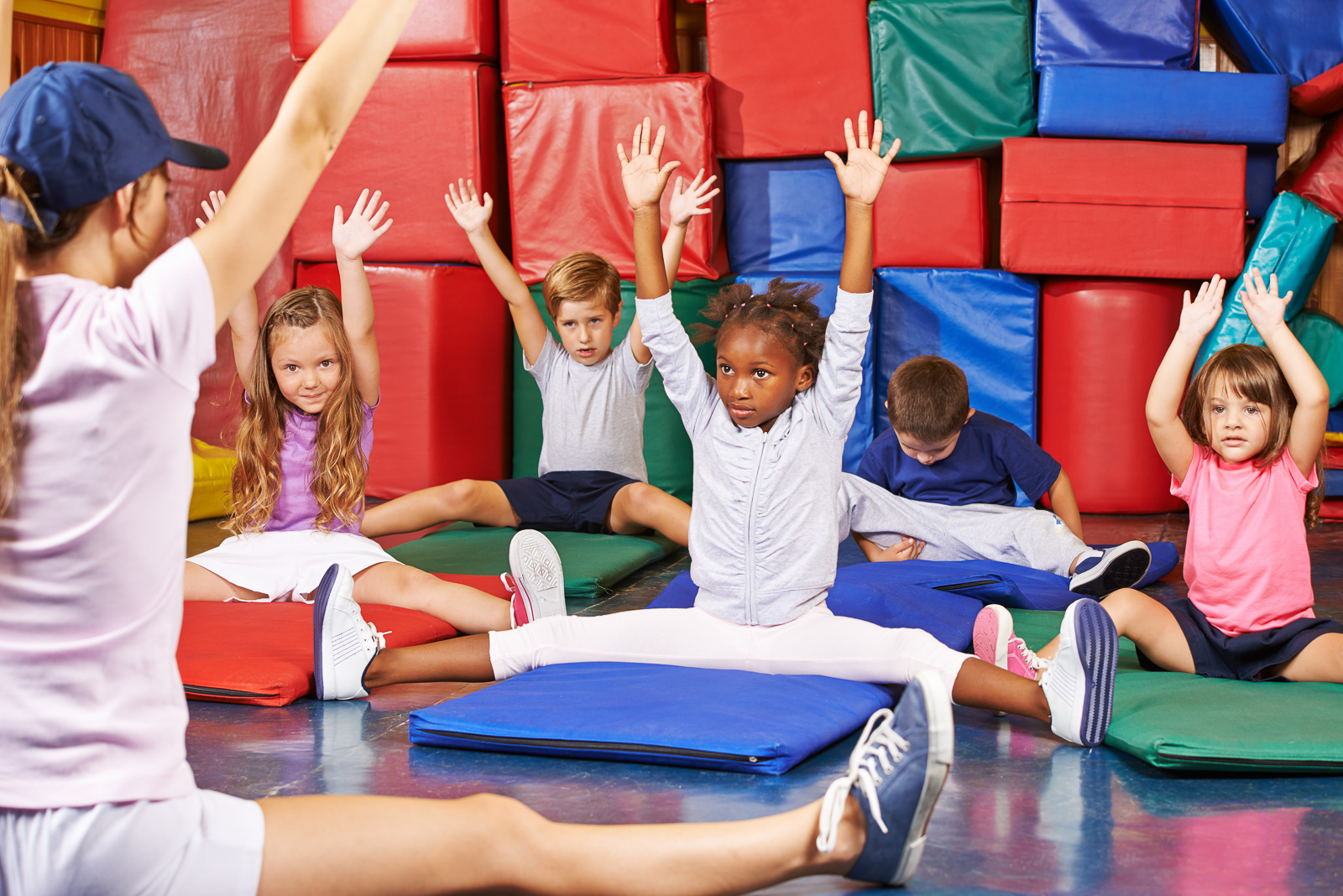 PE with young children – pupils in front of a colourful wall sitting with their arms in the air