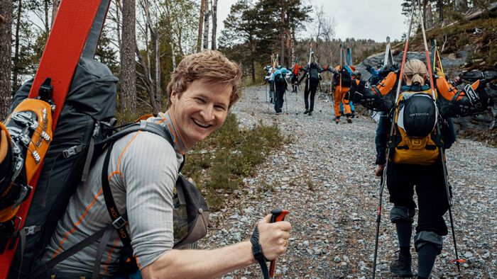 Mann som går med stor sekk og ski på ryggen sammen med en stor gruppe, smiler til kamera mens de går opp en bakke. 