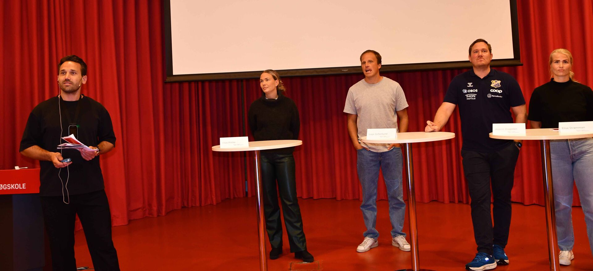 Christian Thue Bjølrndal og fire paneldeltakere står  ved høye cafebord på podiet