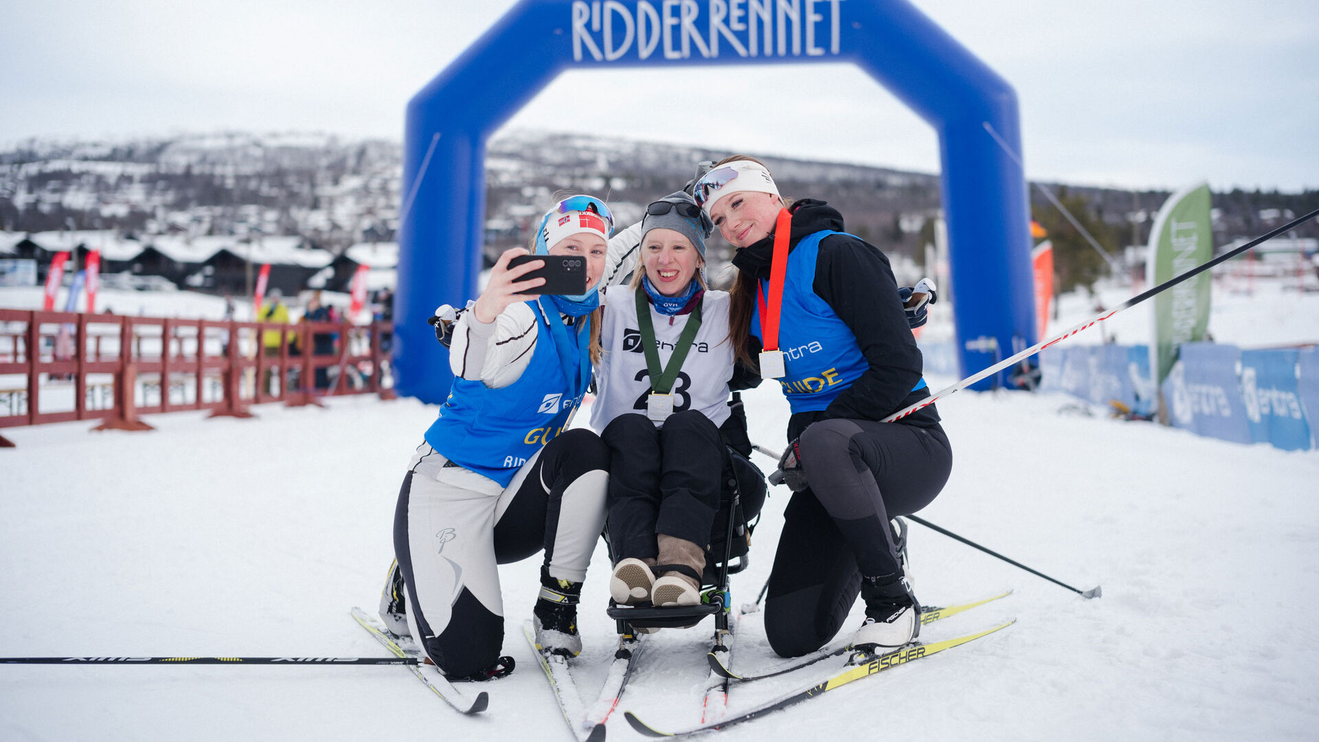 To studenter tar en selfie med en deltaker på sitski med Ridderuke-banner bak