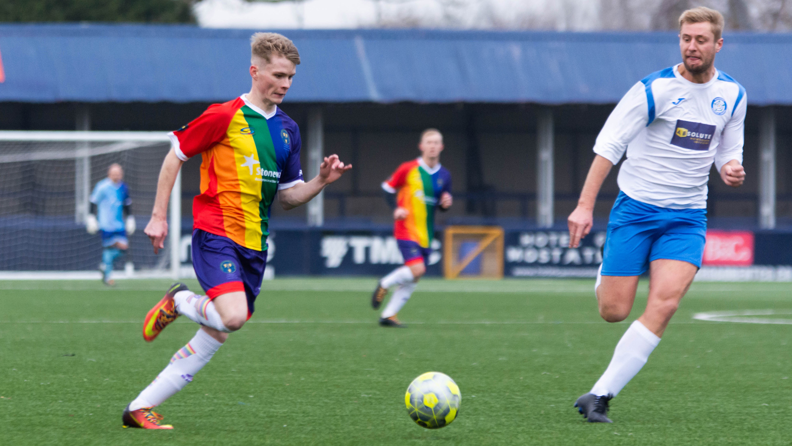 Fotballspillere som løper mot ball. Den ene spilleren har regnbuedrakt.